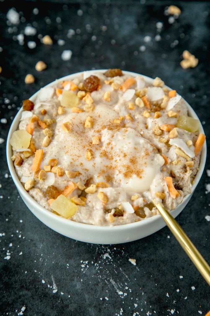 A white bowl filled with keto carrot cake oatmeal and topped with shredded carrots, walnuts, raisins, dried pineapple and a pool of coconut butter frosting.