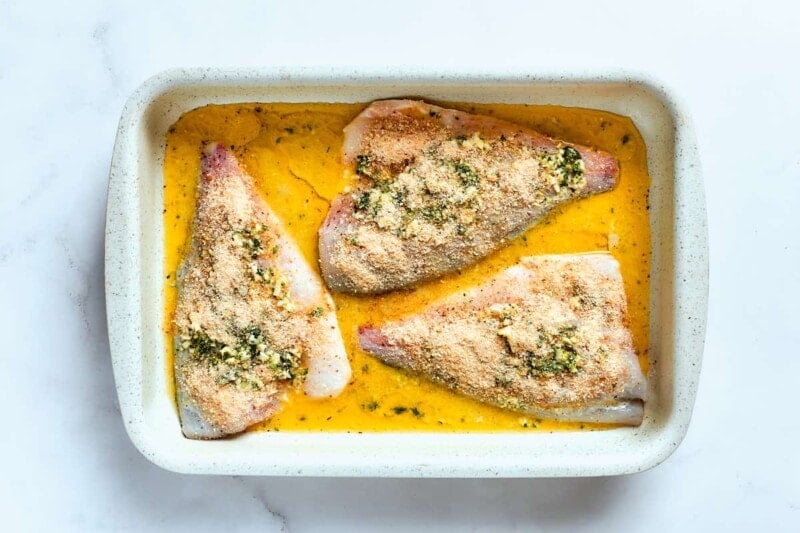 raw seasoned flounder fillets in baking dish with garlic butter and bread crumbs on top.