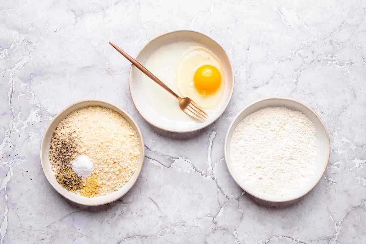 three bowls filled with seasonings and panko in one, egg in another, and flour in the third.