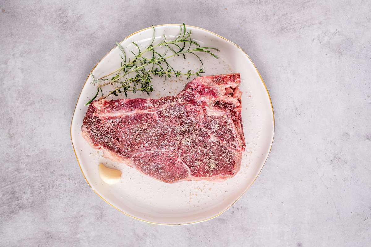 seasoned raw steak on a plate with garlic and Rosemary.