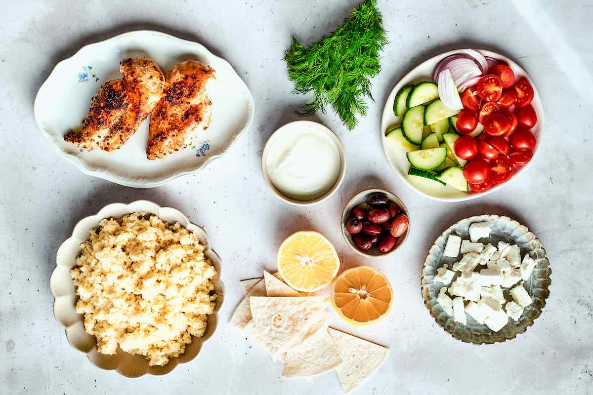 assembling chicken bowls with Greek chicken, vegetables, couscous, and feta.