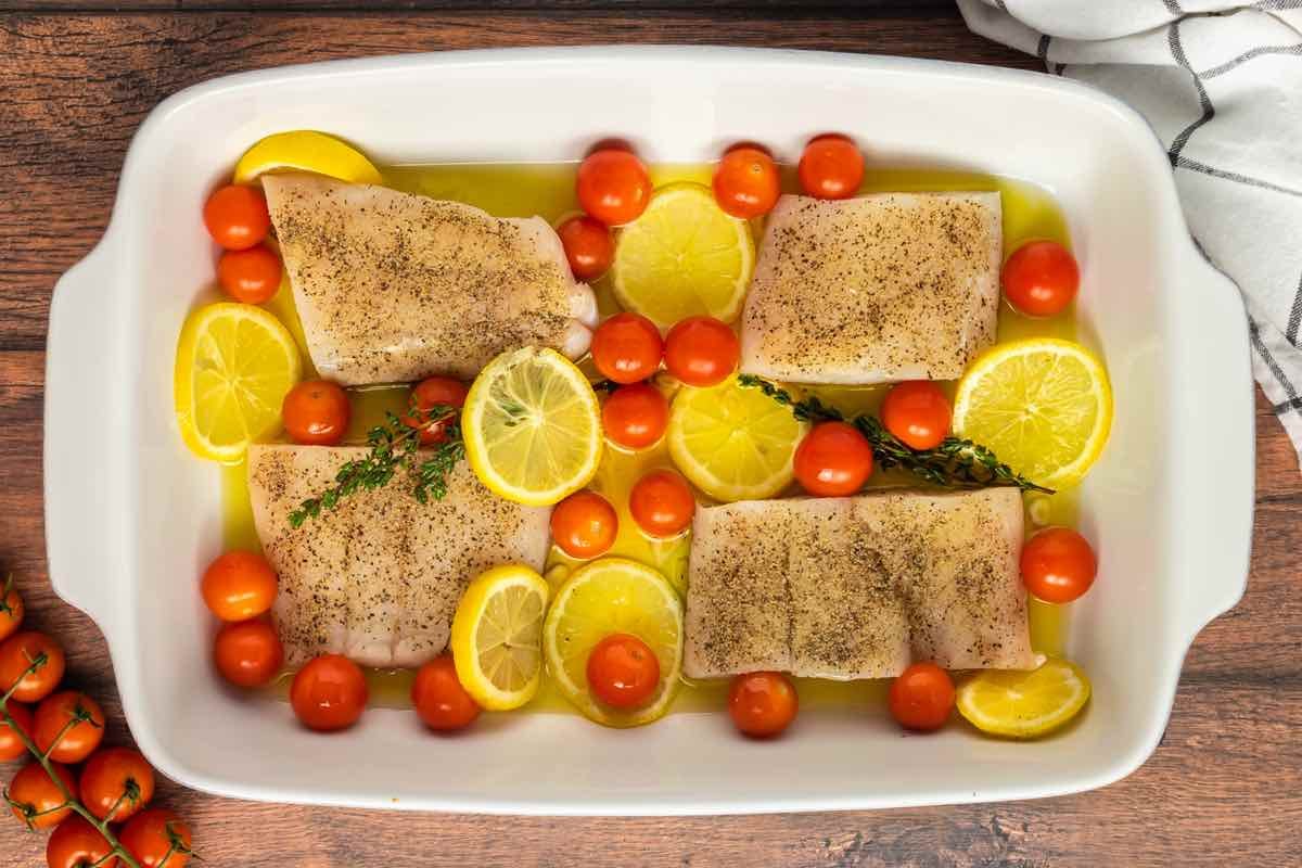 olive oil, tomatoes, and lemons in a baking dish with seasoned raw haddock fillets on top.