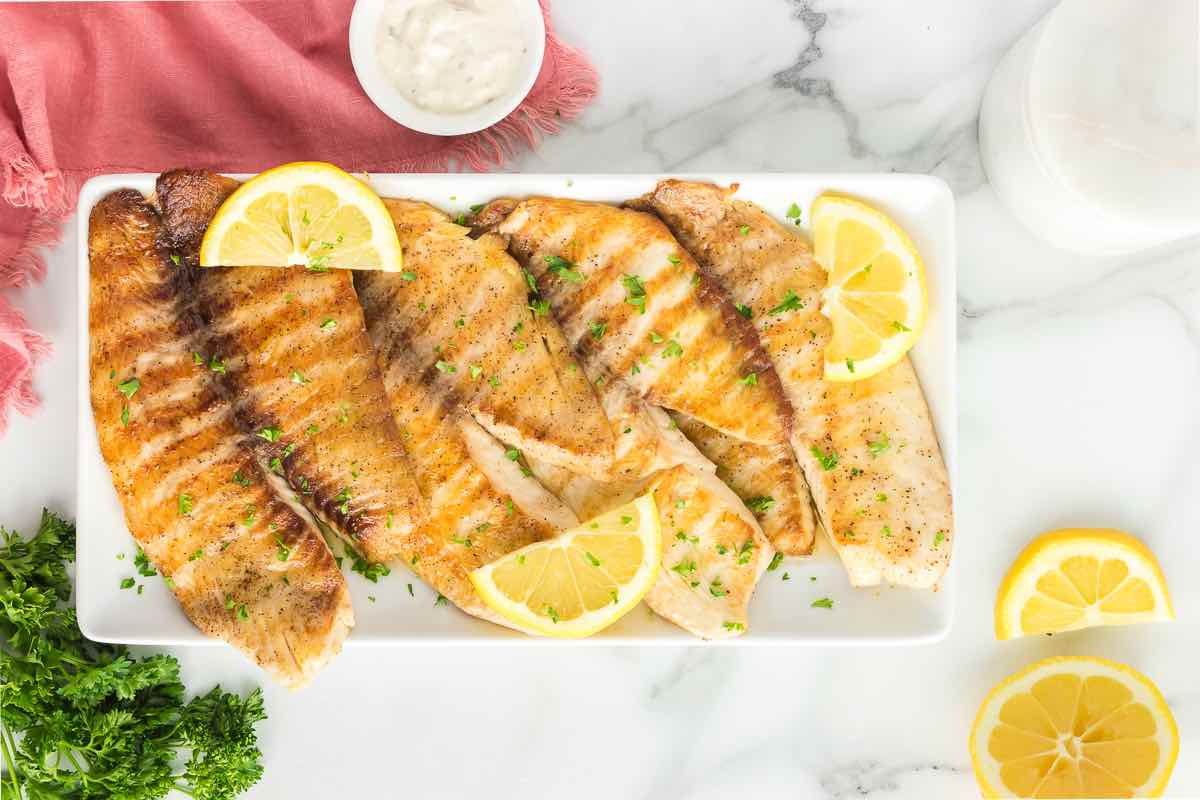 grilled tilapia fillets on a plate with sliced lemons and herbs.