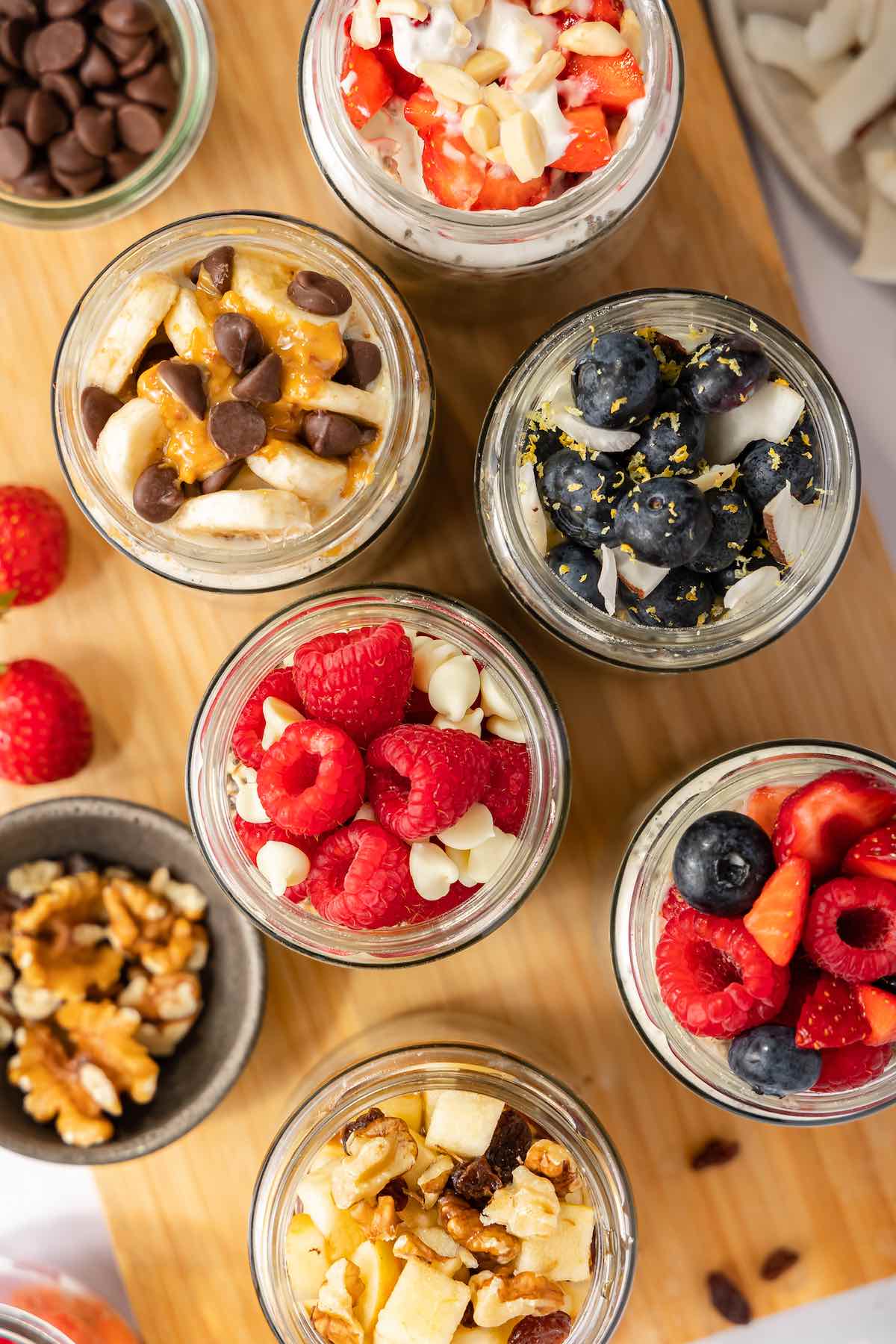 Overhead view of six jars of protein overnight oats with toppings including raspberries and white chocolate, blueberries and coconut, mixed berries, apple with pecans and raisins, strawberries and almonds, and banana with peanut butter and chocolate chips.