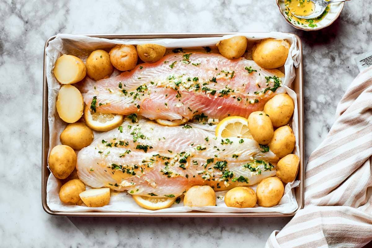 raw, seasoned pollock fillets and raw potatoes on a baking dish.