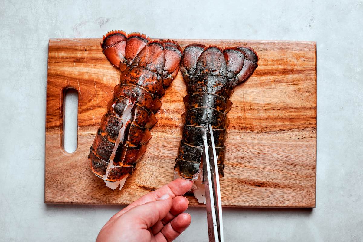 Two lobster tails on a chopping board with scissors cutting into one of them.