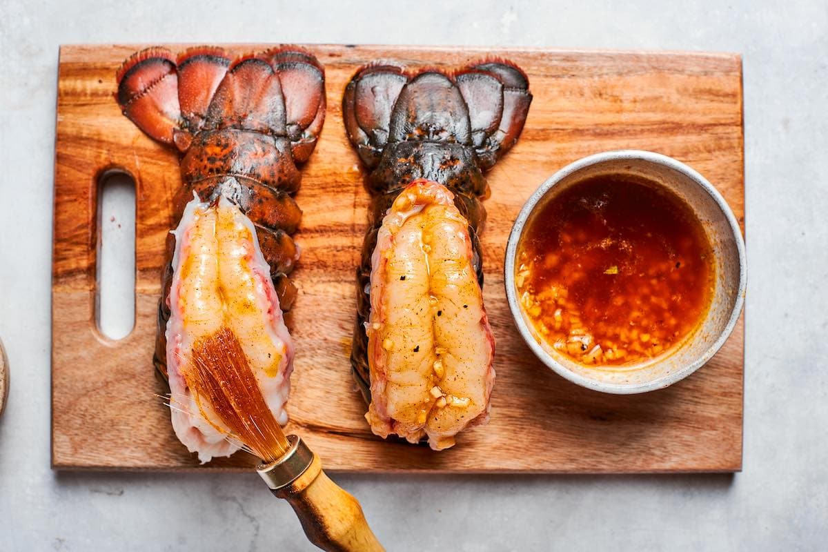 two lobster tails on a chopping board with garlic butter brushed on top.
