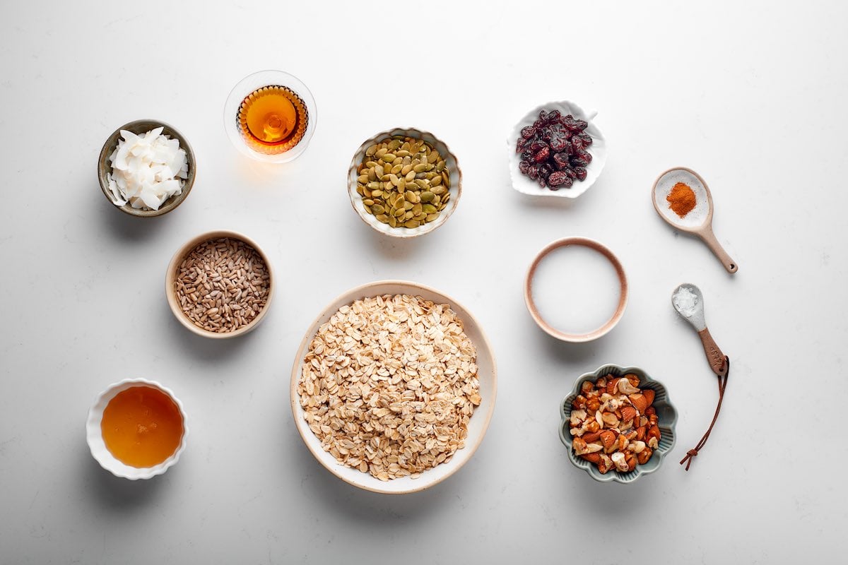 ingredients for air fryer granola: a bowl of oats, chopped nuts, seeds, maple syrup, honey, cranberries, and shredded coconut.