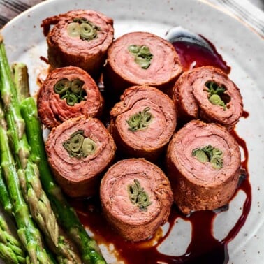 Beef negimaki rolls sliced to show scallion centers on a white plate with sauce and asparagus.
