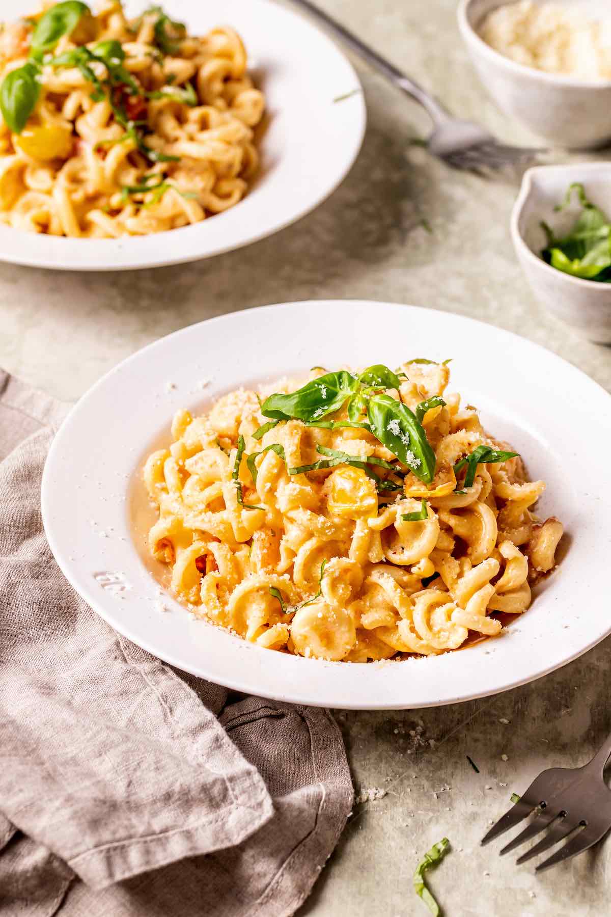 Two white bowls of Boursin cheese pasta with cavatappi, creamy tomato sauce, fresh basil leaves, and Parmesan with a linen napkin.