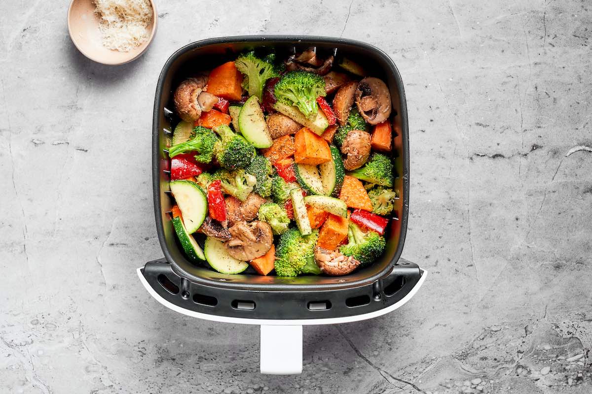 Raw mixed vegetables including broccoli, sweet potato, zucchini, mushrooms, and red bell pepper in an air fryer basket before cooking.