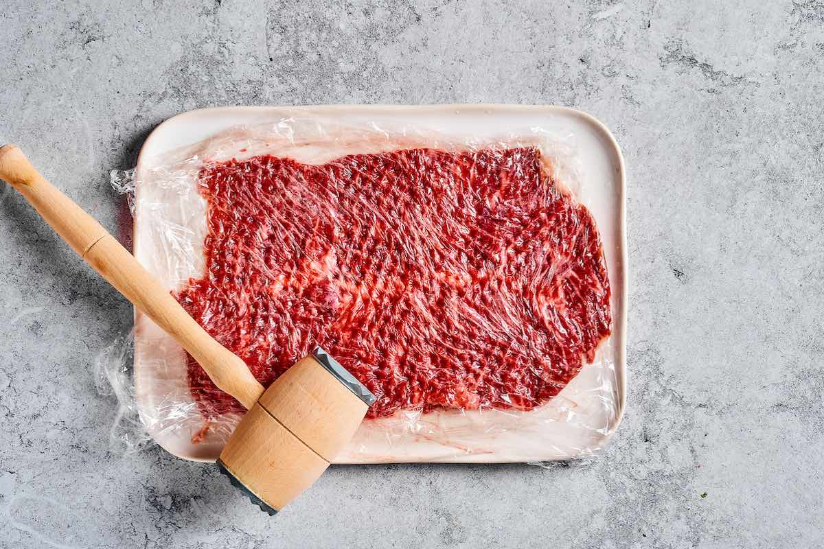 Flank steak being pounded thin between plastic wrap with a meat mallet for beef negimaki.