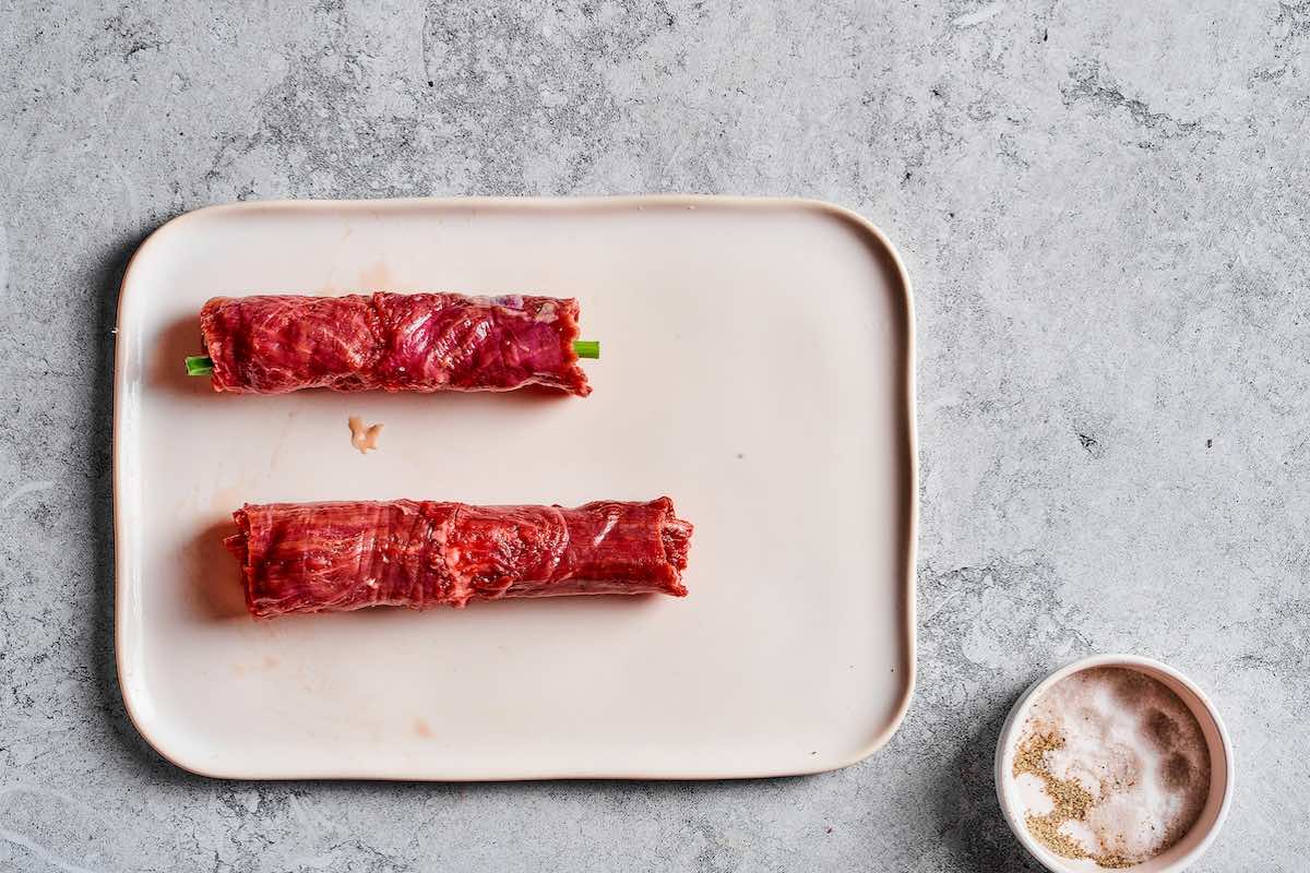 Two raw beef negimaki rolls with scallions on a white plate before cooking.