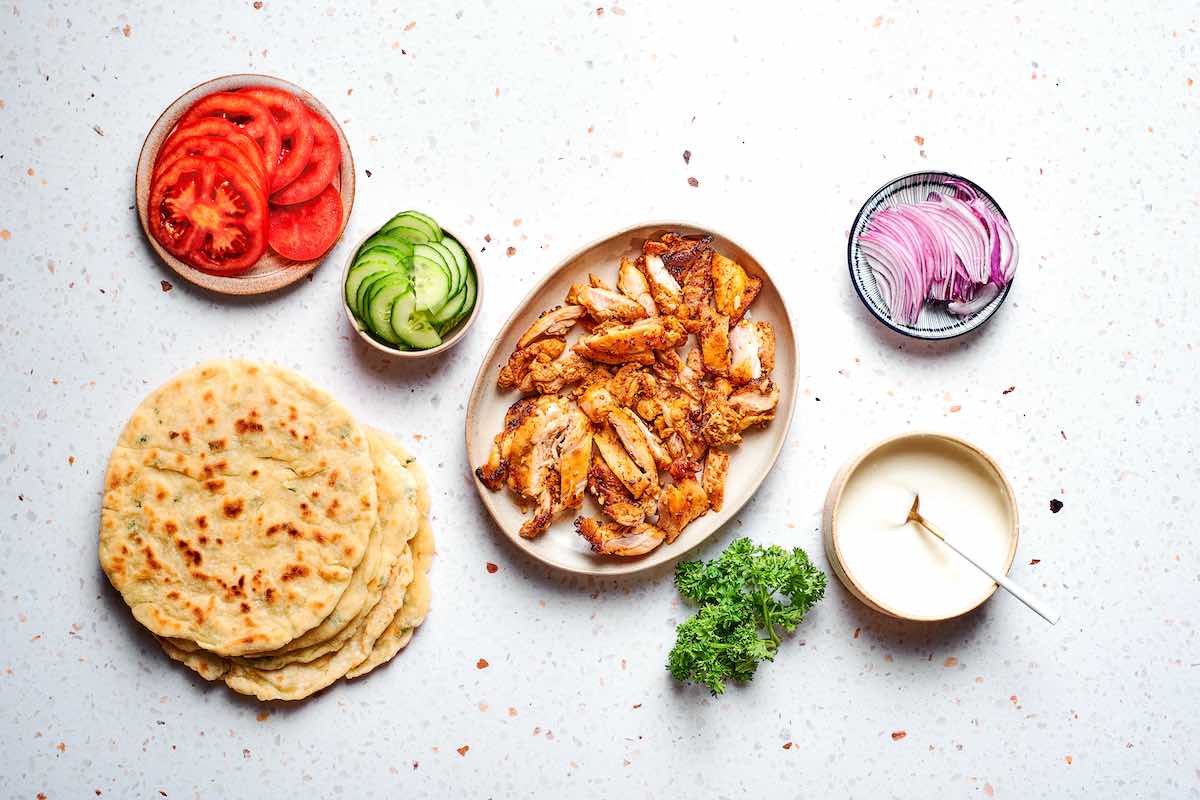 sliced air fried chicken shawarma on a plate with pita, tomatoes, red onion, parsley, cucumber, and yogurt sauce.