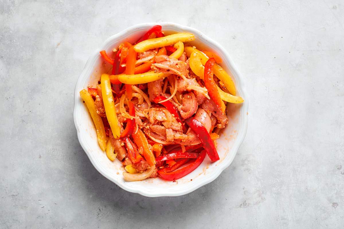 raw chicken, bell peppers, and onion in a mixing bowl, coated in fajita seasoning mix.